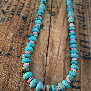 May include: A turquoise and pink beaded necklace with gold accents. The beads are round and have a marbled pattern. The necklace is lying on a wooden surface.