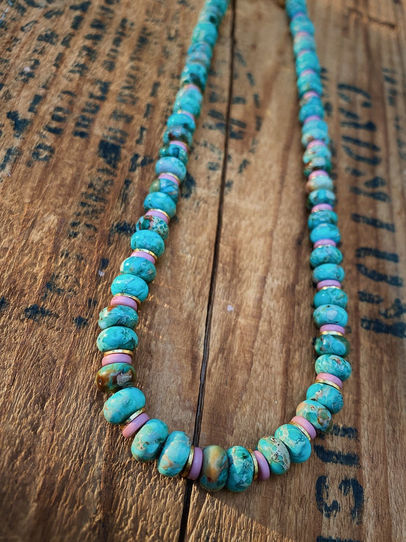 May include: A turquoise and pink beaded necklace with gold accents. The beads are round and have a marbled pattern. The necklace is lying on a wooden surface.