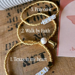 May include: Three gold beaded bracelets. One bracelet has the text "Texas" on white letter beads, another says "Walk by Faith", and the third says "Pray on it". Each bracelet has a unique charm.