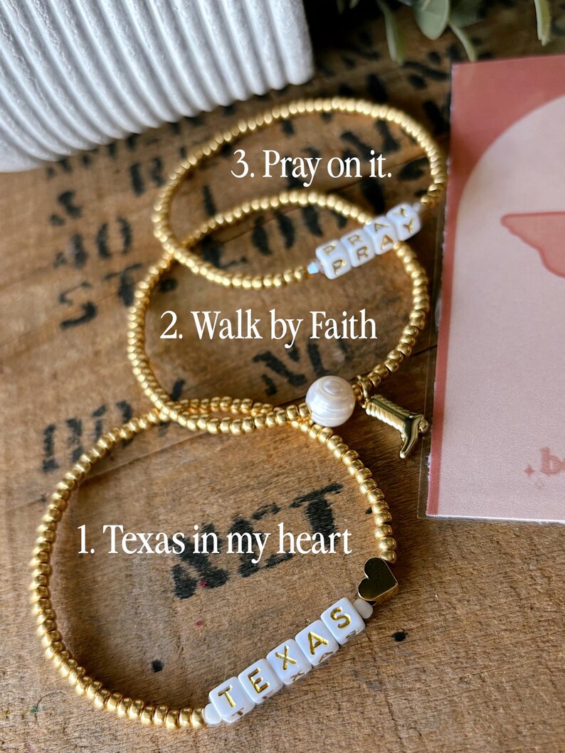May include: Three gold beaded bracelets. One bracelet has the text "Texas" on white letter beads, another says "Walk by Faith", and the third says "Pray on it". Each bracelet has a unique charm.