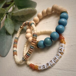 Puede incluir: Tres pulseras de cuentas con un esquema de colores blanco, azul y naranja. Las pulseras est&aacute;n apiladas una encima de la otra. La pulsera inferior tiene cuentas blancas con las palabras "WORTH THE" escritas en letras doradas. La pulsera del medio tiene cuentas blancas con acentos azules y marrones. La pulsera superior tiene cuentas de madera.