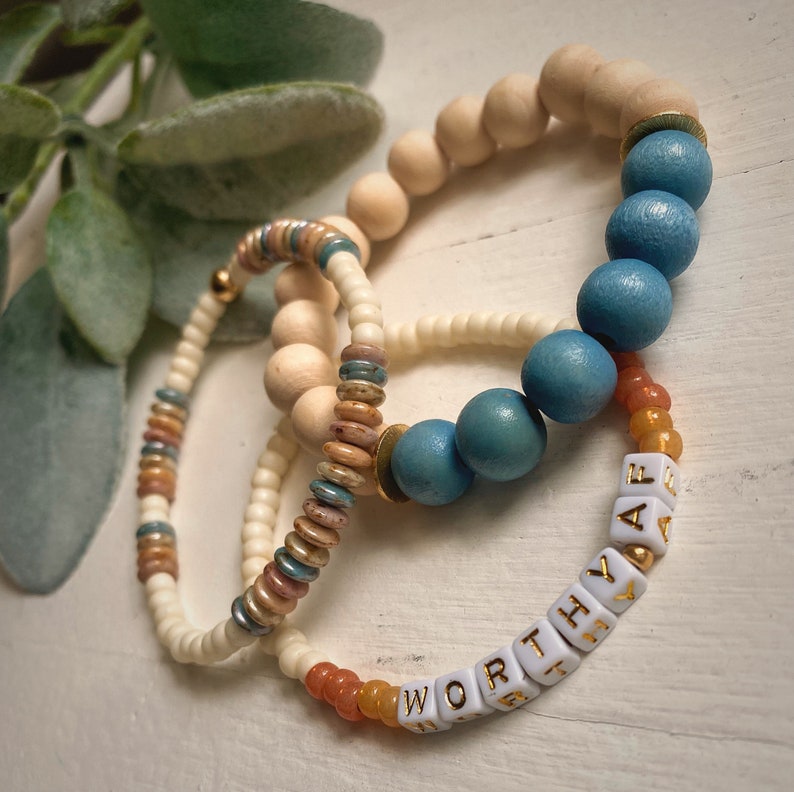 Puede incluir: Tres pulseras de cuentas con un esquema de colores blanco, azul y naranja. Las pulseras est&aacute;n apiladas una encima de la otra. La pulsera inferior tiene cuentas blancas con las palabras "WORTH THE" escritas en letras doradas. La pulsera del medio tiene cuentas blancas con acentos azules y marrones. La pulsera superior tiene cuentas de madera.