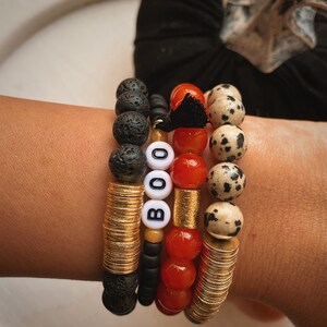Puede incluir: Pulseras apiladas con roca volcánica, discos dorados, cuentas rojas y cuentas de jaspe dálmata. Una pulsera deletrea "BOO" con cuentas de letras blancas. Una borla negra añade un toque decorativo.