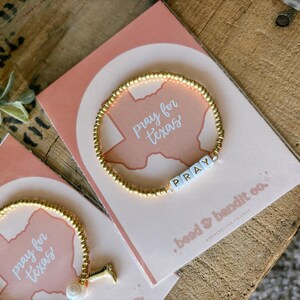 May include: Gold beaded bracelet with the word "PRAY" spelled out in white letter beads. The bracelet is displayed on a pink card with the words "pray for texas" and a Texas outline. The card is on a wooden surface.