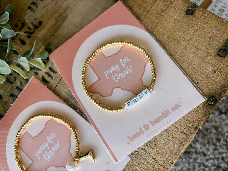 May include: Gold beaded bracelet with the word "PRAY" spelled out in white letter beads. The bracelet is displayed on a pink card with the words "pray for texas" and a Texas outline. The card is on a wooden surface.