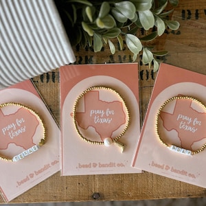 May include: Gold beaded bracelets with the words "TEXAS" and "PRAY" on white letter beads. Each bracelet is on a pink card with a Texas outline and the words "pray for texas". The cards are from "bead & bandit co."
