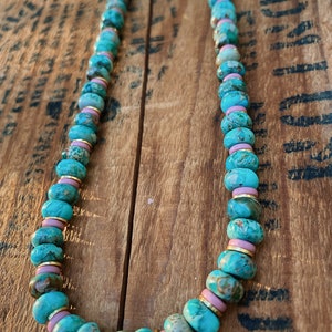 May include: A turquoise and pink beaded necklace with gold accents. The beads are round and have a marbled pattern. The necklace is on a wooden surface.