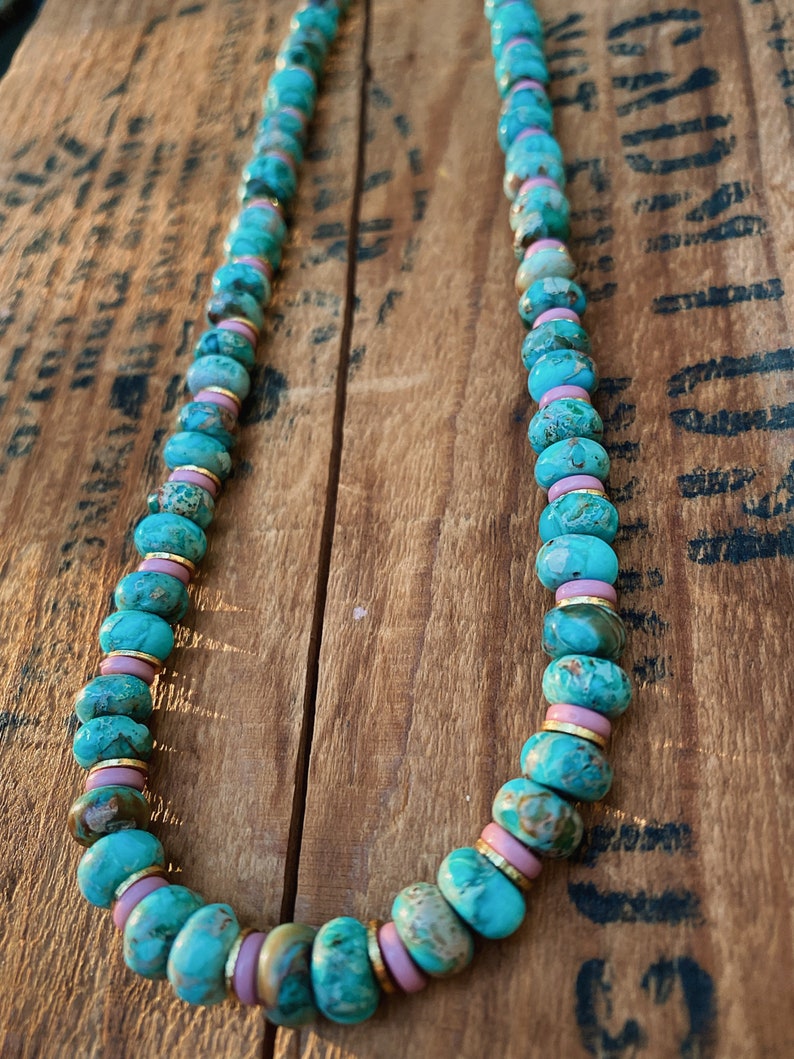 May include: A turquoise and pink beaded necklace with gold accents. The beads are round and have a marbled pattern. The necklace is on a wooden surface.