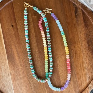 May include: Three beaded necklaces with gold clasps. The necklaces are made of different colored stones, including turquoise, pink, yellow, and green. The necklaces are arranged in a rainbow pattern.