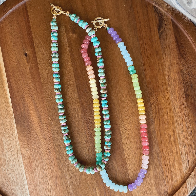 May include: Three beaded necklaces with gold clasps. The necklaces are made of different colored stones, including turquoise, pink, yellow, and green. The necklaces are arranged in a rainbow pattern.