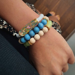 Puede incluir: Un conjunto de tres pulseras con cuentas azules, blancas y verdes. La pulsera superior tiene un tubo transparente con remolinos coloridos en su interior.
