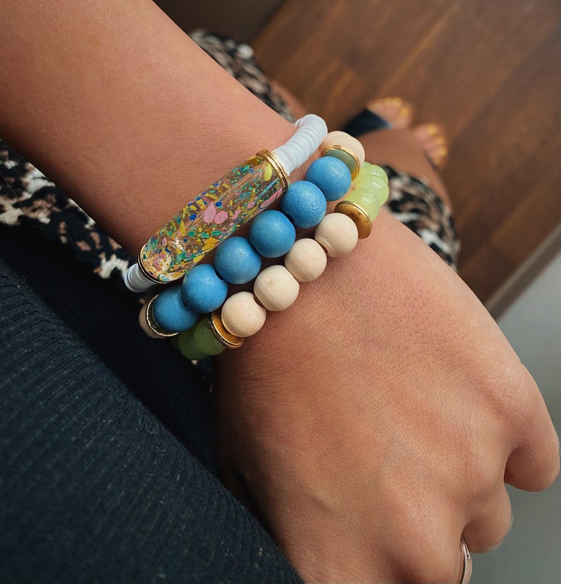 Puede incluir: Un conjunto de tres pulseras con cuentas azules, blancas y verdes. La pulsera superior tiene un tubo transparente con remolinos coloridos en su interior.