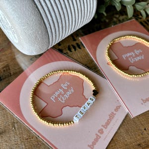 May include: Gold beaded bracelets with the word "TEXAS" spelled out in white letter beads. The bracelets are on pink cards with the words "pray for texas" and a Texas outline. The background is a wooden surface.