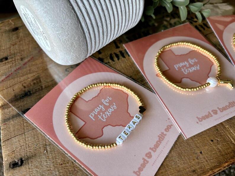May include: Gold beaded bracelets with the word "TEXAS" spelled out in white letter beads. The bracelets are on pink cards with the words "pray for texas" and a Texas outline. The background is a wooden surface.