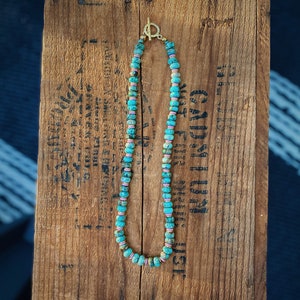 May include: A turquoise and pink beaded necklace with a gold clasp. The necklace is on a wooden surface.