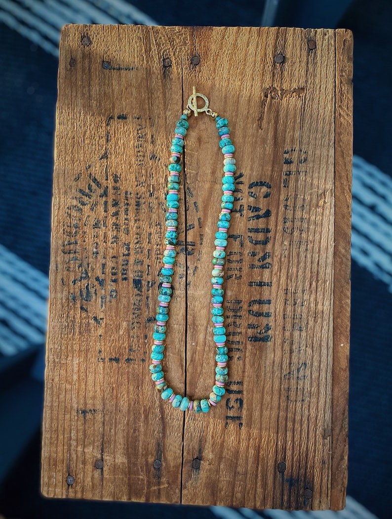 May include: A turquoise and pink beaded necklace with a gold clasp. The necklace is on a wooden surface.
