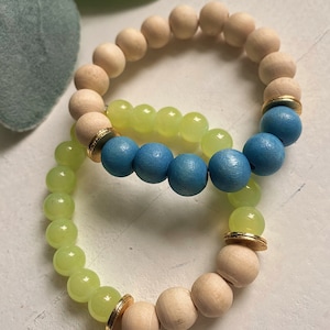 Puede incluir: Dos pulseras hechas de cuentas de madera, una con cuentas de color azul claro y la otra con cuentas de color verde claro. Ambas pulseras tienen detalles dorados.