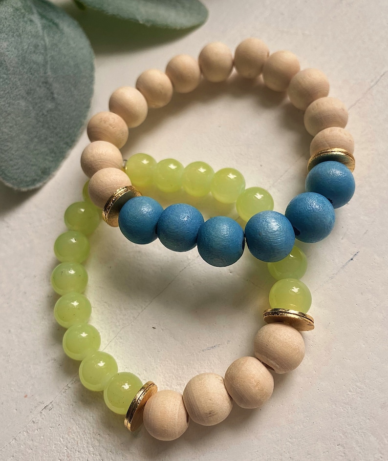 Puede incluir: Dos pulseras hechas de cuentas de madera, una con cuentas de color azul claro y la otra con cuentas de color verde claro. Ambas pulseras tienen detalles dorados.