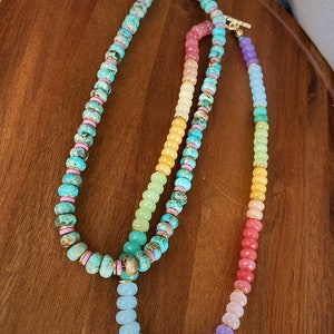 May include: Two layered necklaces with colorful gemstone beads. The top necklace has turquoise, pink, and yellow beads. The bottom necklace has pink, yellow, green, blue, and purple beads.