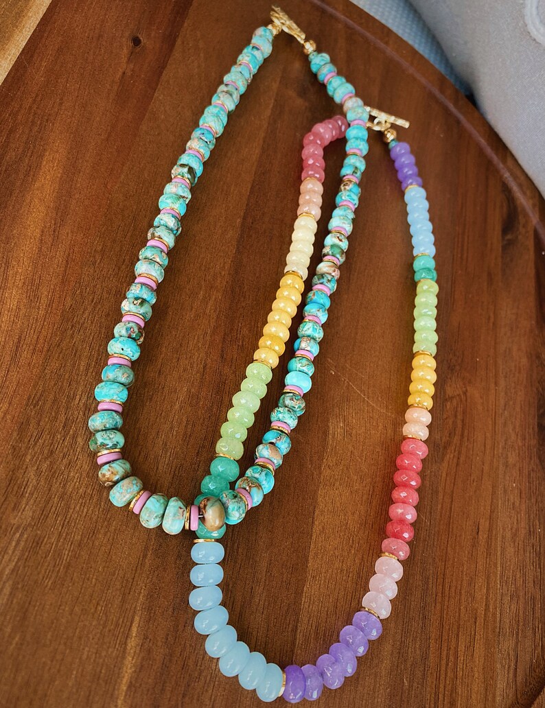 May include: Two layered necklaces with colorful gemstone beads. The top necklace has turquoise, pink, and yellow beads. The bottom necklace has pink, yellow, green, blue, and purple beads.
