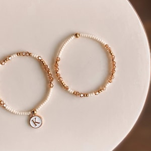 May include: Two beaded bracelets. One bracelet features a white band with gold-coloured beads and a charm with the letter "K". The other bracelet has a similar design. The bracelets are on a white surface.