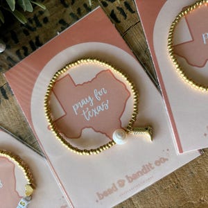 May include: Gold beaded bracelets with charms and the words "pray for texas" on cards. One bracelet has a pearl and a charm, another has letter beads spelling "PR". The cards have a Texas outline.