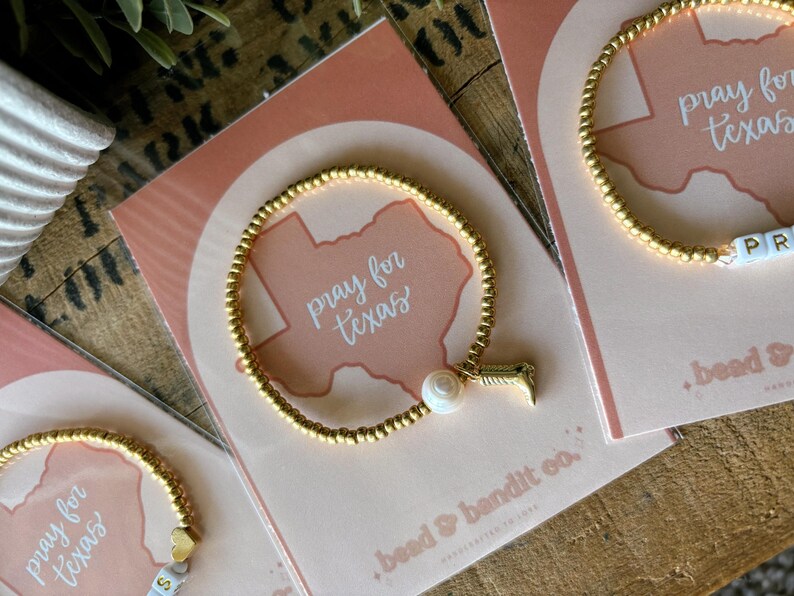 May include: Gold beaded bracelets with charms and the words "pray for texas" on cards. One bracelet has a pearl and a charm, another has letter beads spelling "PR". The cards have a Texas outline.