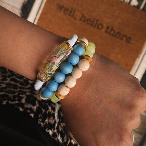 Puede incluir: Un conjunto de tres pulseras que incluyen una pulsera de resina colorida con purpurina, una pulsera de cuentas de madera azul claro y una pulsera de cuentas verde claro y doradas.