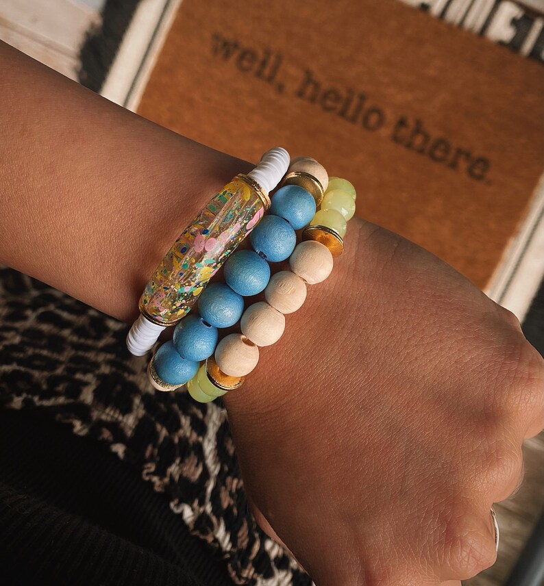 Puede incluir: Un conjunto de tres pulseras que incluyen una pulsera de resina colorida con purpurina, una pulsera de cuentas de madera azul claro y una pulsera de cuentas verde claro y doradas.