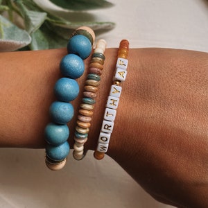 Puede incluir: Tres pulseras apiladas en una mu&ntilde;eca. La pulsera superior est&aacute; hecha de peque&ntilde;as cuentas blancas con letras doradas que deletrean "WORTHY AF". La pulsera del medio est&aacute; hecha de peque&ntilde;as cuentas marrones, blancas y azules. La pulsera inferior est&aacute; hecha de cuentas azules grandes con un acento dorado.