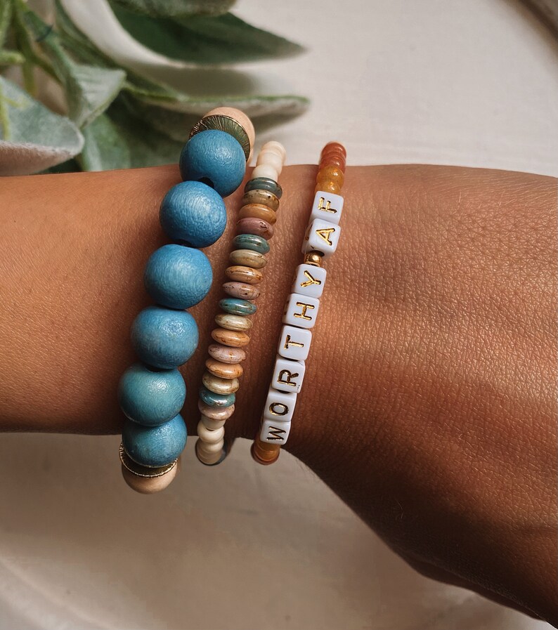 Puede incluir: Tres pulseras apiladas en una mu&ntilde;eca. La pulsera superior est&aacute; hecha de peque&ntilde;as cuentas blancas con letras doradas que deletrean "WORTHY AF". La pulsera del medio est&aacute; hecha de peque&ntilde;as cuentas marrones, blancas y azules. La pulsera inferior est&aacute; hecha de cuentas azules grandes con un acento dorado.