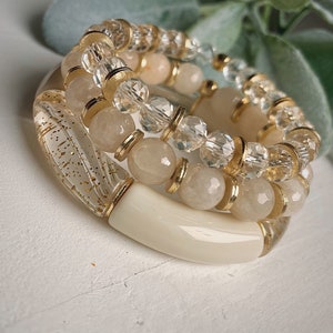 May include: Three stacked bracelets with gold accents. The bottom bracelet is white and has a textured surface. The middle bracelet is made of clear and opaque white beads with gold accents. The top bracelet is made of clear beads with gold accents.