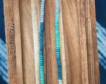 May include: A graduated ombre necklace made of light blue, teal, and green gemstone beads. The necklace has a gold clasp.