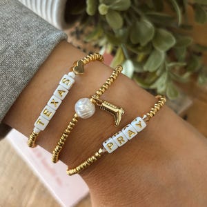 May include: Three gold beaded bracelets. One bracelet spells "TEXAS" with white letter beads. Another has a pearl and a gold cowboy boot charm. The third bracelet spells "PRAY" with white letter beads.