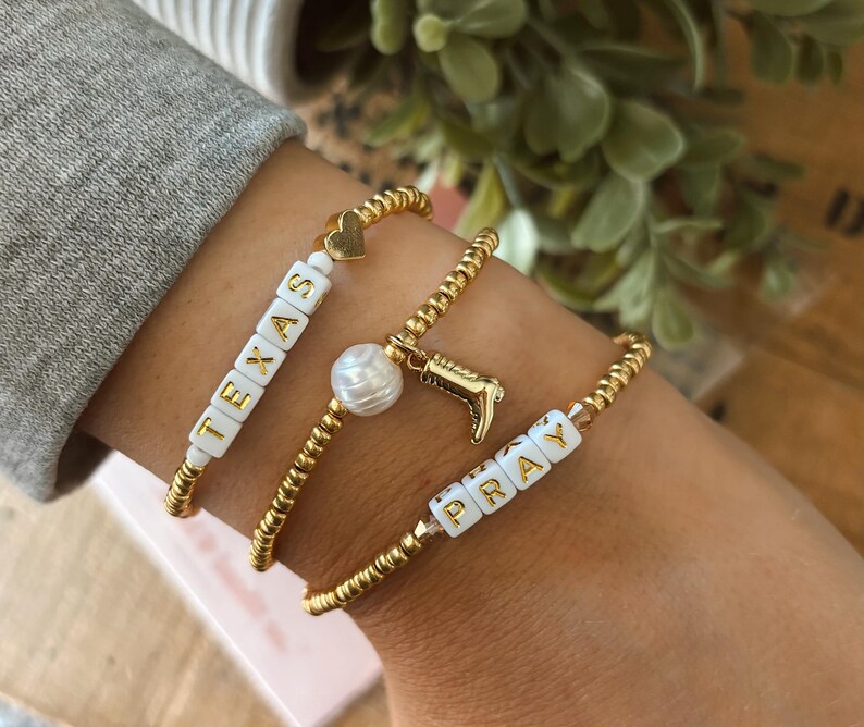 May include: Three gold beaded bracelets. One bracelet spells "TEXAS" with white letter beads. Another has a pearl and a gold cowboy boot charm. The third bracelet spells "PRAY" with white letter beads.