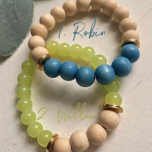 Puede incluir: Dos pulseras de cuentas, una con cuentas azul claro y amarillas, la otra con cuentas verde claro y de madera natural. Las pulseras est&aacute;n etiquetadas como "1. Robin" y "2. Willow".