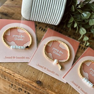 May include: Gold beaded bracelets with white lettered beads spelling "TEXAS" and "PRAY". Each bracelet is displayed on a pink card with the text "pray for texas" and "bead & bandit co."