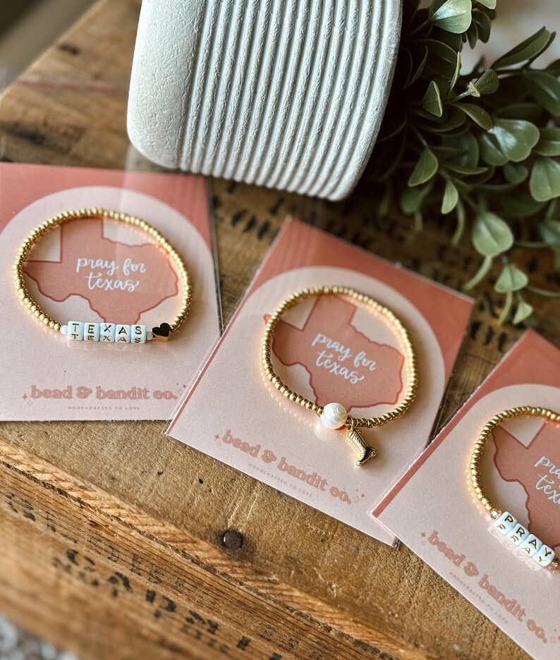 May include: Gold beaded bracelets with white lettered beads spelling "TEXAS" and "PRAY". Each bracelet is displayed on a pink card with the text "pray for texas" and "bead & bandit co."