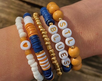 May include: A set of five bracelets featuring a heart charm, white and orange striped beads, blue and white beads, gold beads, and white beads with the words "STL" and "BLUES" in gold lettering.