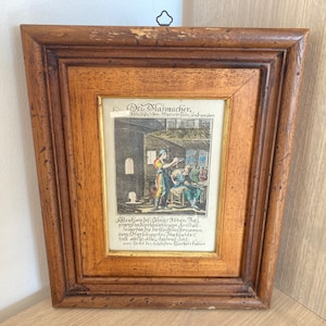 May include: A framed antique print of a glassblower at work. The print is in a wooden frame with a gold-colored mat. The text on the print reads "Der Glasmacher,  Vorhin des Herrn von der Schulenburg."  The text below the image reads "Allemone, die Slate, Richen, Sal,  prungene, die,  verkaufft,  und  verwandelt,  bey  der  Glas-Hutte,  bey  der  Oeffentlichen  Schreiberey,  und  auf  dem  alten  Marckt  zu  Eimbeck."