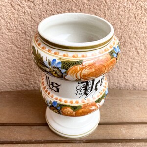 Peut inclure: Un pot en céramique blanche avec un motif floral dans des tons de bleu, vert, orange et marron. Le pot a une large ouverture et une base ronde. Le pot est décoré d'un motif floral et du mot "Piel" en lettres noires.