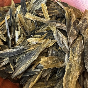 May include: Close-up of dried tea leaves. The leaves are long, thin, and have a mix of dark green and golden-brown hues. The texture appears rough and natural, suggesting a high-quality, unprocessed product. The leaves are piled together, filling the frame.