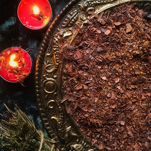 May include: An overhead shot features a decorative, antique-style brass tray filled with reddish-brown tea leaves. Two lit red candles with floral accents sit nearby. A bundle of dried herbs and a yellow crystal are also present, creating a mystical aesthetic.