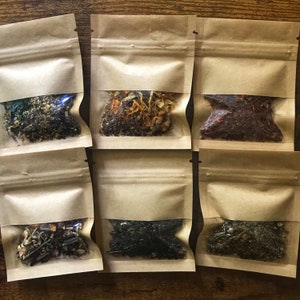 May include: Six small, clear-windowed pouches filled with loose leaf tea. The pouches are sealed and made of brown paper. The tea varieties include green, black, and herbal blends, with visible flowers and herbs.