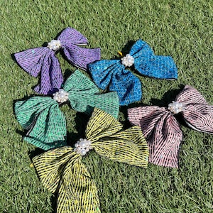 Puede incluir: Seis lazos de pelo de tela brillante en varios colores, cada uno con un acento de perla. Los lazos están dispuestos sobre una superficie de hierba verde.