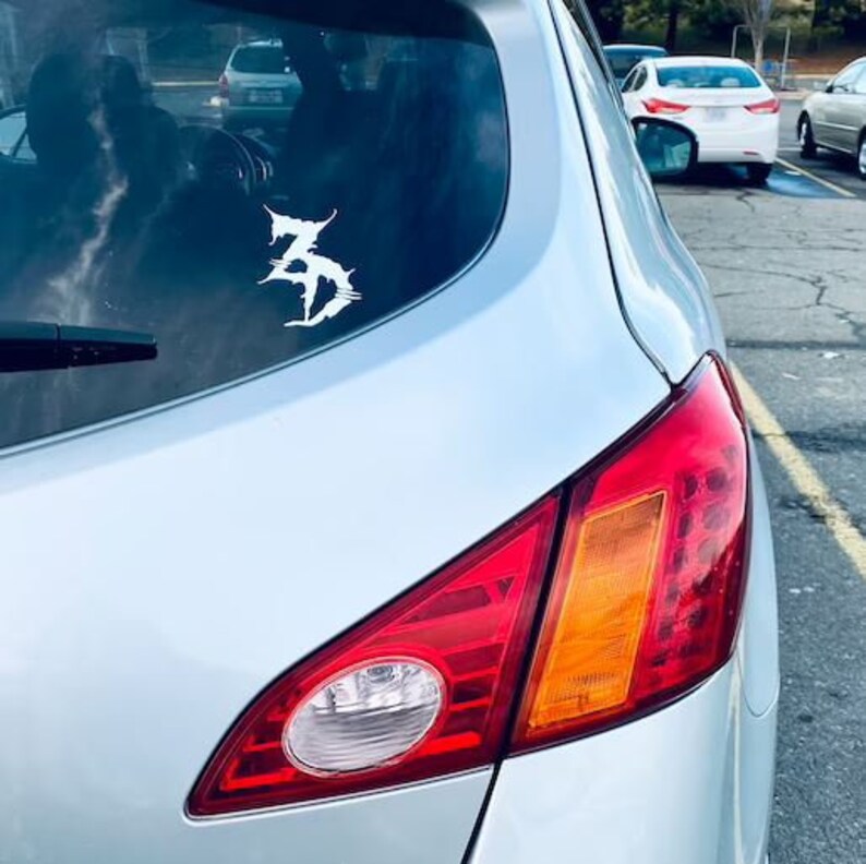 May include: A silver car with a white sticker of a stylised letter Z on the rear window. The car has a red rear light with an orange indicator.