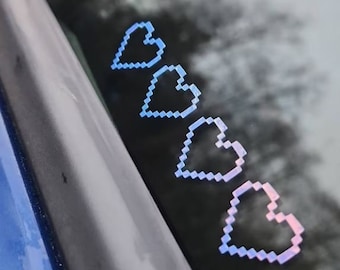 May include: Four pixelated heart stickers in shades of blue and pink are displayed on a window. The hearts are arranged diagonally, with the colors transitioning from blue to pink. The stickers have a retro, digital aesthetic.