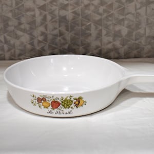 Peut inclure: Une poêle en céramique blanche avec un manche, ornée d'une illustration botanique colorée de légumes et d'herbes sur le côté extérieur. Sous le motif, le texte Le Persil est imprimé en écriture cursive noire sur la surface blanche lisse.