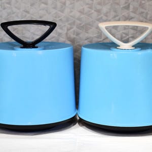 May include: Two light blue, cylindrical containers with black bases and triangular handles. One has a black handle, the other a cream handle. The containers are on a white surface against a textured grey background.
