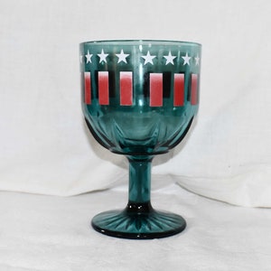 May include: A teal glass goblet with a white star and red stripe design. The goblet has a wide bowl and a tall stem.